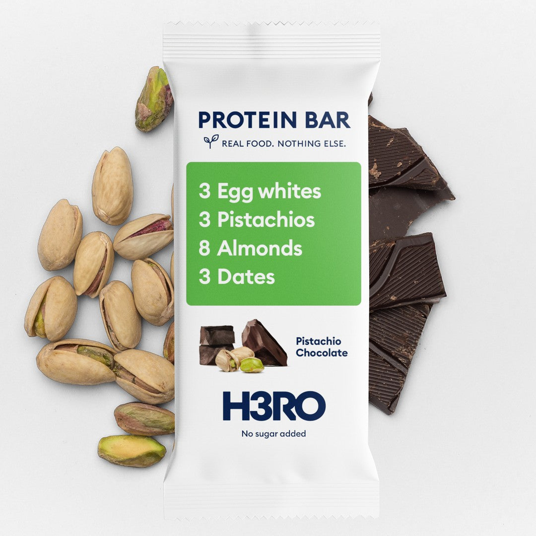 A white H3RO protein bar package labeled Pistachio Chocolate, surrounded by whole pistachios, dark chocolate chunks, and a green ingredient panel showing egg whites, pistachios, almonds, and dates on a light gray background.